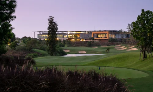 View of the Nikanti Golf Club course at dusk, showcasing a modern clubhouse in the background. The foreground features a well-manicured green with a tee box and multiple strategically placed bunkers. The fairway is bordered by trees and bushes, with a water hazard adding to the challenge. The lush, rolling terrain and pristine landscaping highlight the course's design, offering golfers a visually stunning and challenging round.