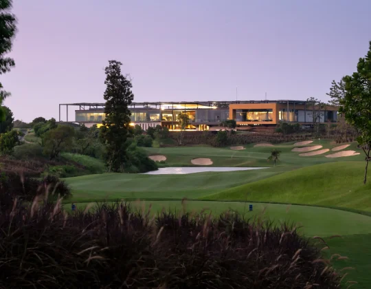 View of the Nikanti Golf Club course at dusk, showcasing a modern clubhouse in the background. The foreground features a well-manicured green with a tee box and multiple strategically placed bunkers. The fairway is bordered by trees and bushes, with a water hazard adding to the challenge. The lush, rolling terrain and pristine landscaping highlight the course's design, offering golfers a visually stunning and challenging round.